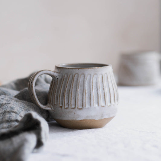 Large hand carved tea mug with a handle in light grey colour. Photographed with a blue tea towel on the left. 